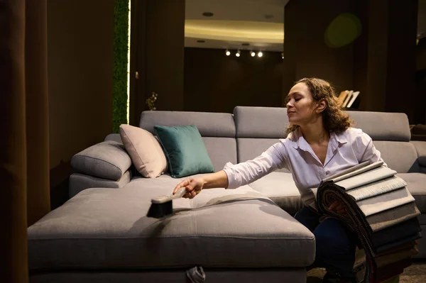 Hispanic creative interior designer, professional decorator holds upholstery fabric samples and cleans the velour surface of a gray art nouveau sofa with the help of a dustpan in the home interior