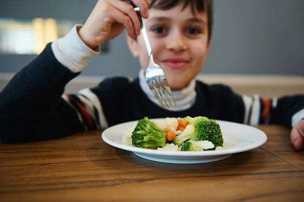 Focus on steamed vegetables, healthy vegan food served in white plate and blurred adorable boy holding fork and smiling looking at camera during lunch in cafeteria