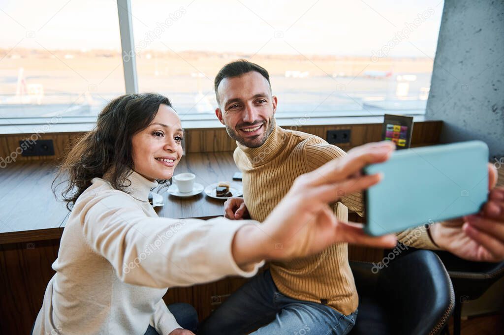 Beautiful multi-ethnic couple in love holding smartphone in outstretched hands and taking self-portrait while enjoying breakfast together in the airport departure terminal before boarding the flight