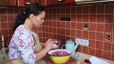 Female chef confectioner, housewife in an apron using a wooden spoon stirs red currants sprinkled with sugar while preparing delicious homemade jelly and preserves in a homely cozy rustic kitchen