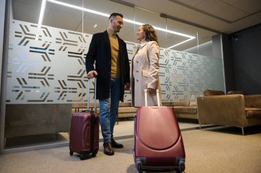 Full length portrait of couple, pretty woman and handsome man with suitcases traveling together, waiting to board a flight in airport VIP lounge. Business trip, team of colleagues, human relationships