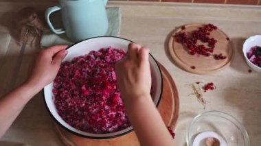 Close-up of stirring red currants with white sugar in a metal enamel bowl in the home kitchen. Vintage blue kettle and red currant berries on a wooden board lying down on a kitchen countertop