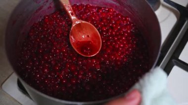 Close-up 4K video of a chef stirring redcurrant berry jam in a metal saucepan on the stove, with a wooden spoon, preparing canned food for the winter season