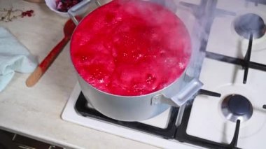 Focus on the metal saucepan on the burning stove with the simmering berry jam and the pastry chef scooping up the froth with a spoon. The stage of making homemade red currant jam.