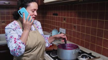 Inspired woman wearing apron, housewife talking on mobile phone while standing at kitchen stove and stirring red currant berry jam with wooden spoon