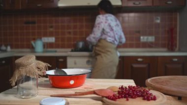 Focus on a red enamel bowl with white dots on a wooden board next to lying, red currant berries against the backdrop of a charming housewife woman standing at the kitchen stove and preparing jam
