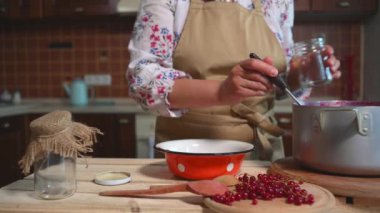 Close-up of a housewife pouring freshly made red currant berry jam into sterilized jars at home rustic kitchen. Jam preservation, bottle sealing, homemade canned food, culinary and cookery concept