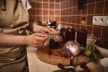 Pasta şefinin ellerinin, yeni yapılmış vişne reçeli kavanozunun kapağına ip bağlaması. Reçel kalıntılarında kirli bir kepçe mutfak atölyesindeki ahşap bir tahtanın üzerinde yatıyor.