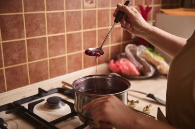 Close-up of the hand of a chef confectioner's holding a kitchen spoon with cherry berries while preparing homemade jam in the saucepan, standing by stove in the home kitchen interior
