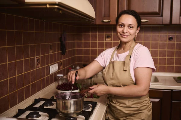 Gorgeous delightful middle-aged multi-ethnic woman, a housewife smiles cutely looking at camera while preparing a homemade cherry jam in the cozy rustic home kitchen. Food and drink concept, culinary