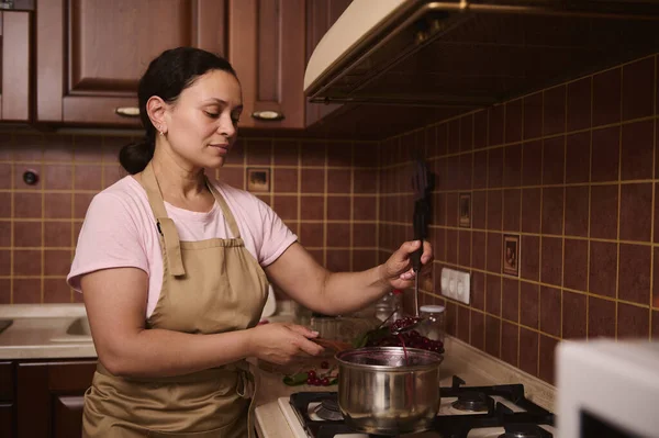 Multiethnic middle-aged woman confectioner, a housewife wearing a chef apron, standing by stove and cooking cherry jam in the pot at home wooden kitchen