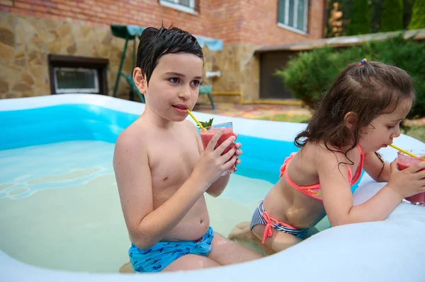 Adorable school age children, Caucasian kids drinking sweet refreshing freshly squeezed fruit cocktail from a straw while having fun in an inflatable swimming pool in the yard of a private house