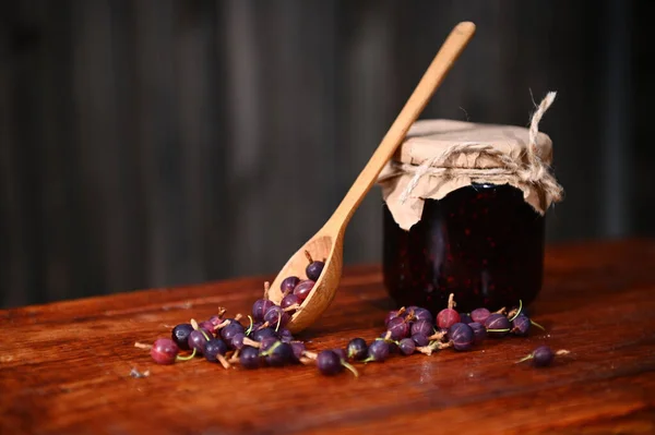 Still life. A glass jar of a homemade wild berry jam, wooden spoon and gooseberries on a rustic wood surface. Canning concept. Sweet healthy food. Organic farm, culinary traditions. Copy space. Banner