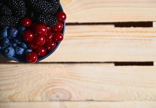 Partial view of fresh organic wild berries - cherries, blackberries, blueberries and bilberries in a blue ceramic bowl in the corner of wooden crate background with advertising space for text. Banner