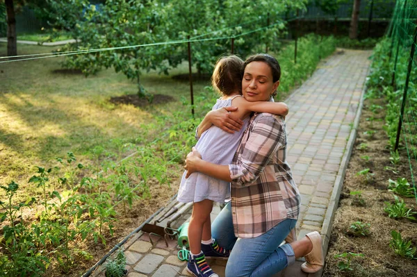 Multiethnic charming woman, loving mother hugging her lovely little daughter, standing together in the eco farm, planting and caring for seedlings. Install love and care for nature since childhood