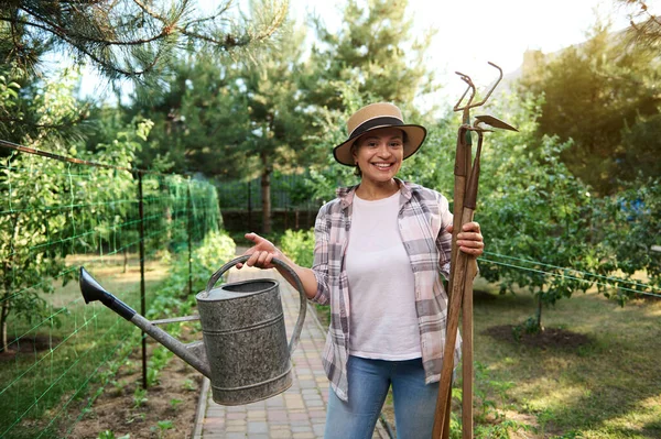 Pleasant female gardener, farmer in a white t-shirt, plaid shirt, blue jeans and a straw hat holds watering can, hoe, ripper and cultivator in hands, in organic vegetable garden, enjoys gardening