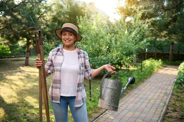Pleasant woman horticulturist holding gardening tools- a watering can, hoe and ripper in her hands, enjoying gardening in organic farm on a sunny early spring day. Beautiful sunbeams on the background