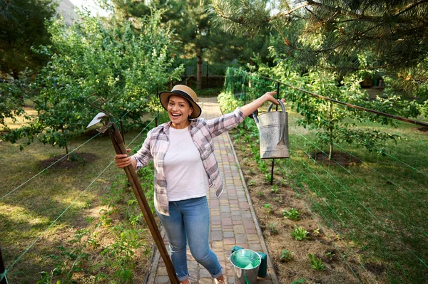 Cheerful female gardener, farmer, horticulturist enjoys gardening on an early spring day, stands in an eco farm with a hoe, ripper and watering can in her hands