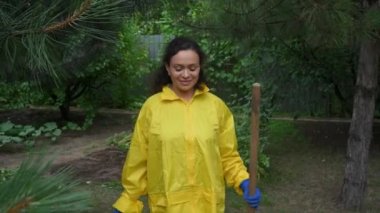 Charming African American woman, farmer in yellow raincoat and blue rubber work gloves walks in the organic farm with a gardening shovel and bucket in her hands. Agriculture, eco farming, horticulture