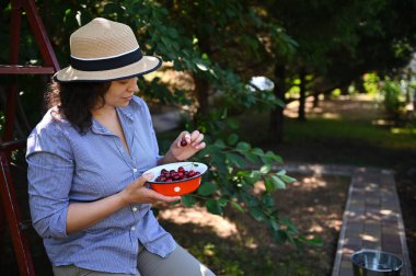 Çekici, koyu renk saçlı kadın, eko çiftçi, kiraz toplayıcısı, hasat şapkası takan, mahsul topladıktan sonra dinlenen, meyve bahçesinde taze toplanmış tatlı kirazlar yiyen. Tarım işi. Çiftçilik