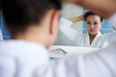 Beyaz bornozlu, siyah saçlı, güzel bir kadına, aynaya yansıyan, banyodaki günlük rutin işlerinde at kuyruğu bağlayan, yumuşak bir odak. Güzellik reklamı. Vücut bakımı