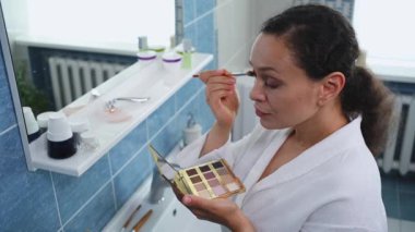 Beautiful middle-aged multiethnic woman in a white bathrobe holds a colorful makeup palette and applies eye shadow while looking at herself in the bathroom mirror. Makeup and appearance care concept