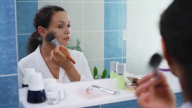 Beyaz bornozlu çekici orta yaşlı kadın makyaj fırçasıyla yüzüne pudra sürüyor, ayna görüntüsüne gülümsüyor, doğal görünüşünden ve makyajından memnun..