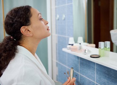 Beyaz bornozlu çekici bir kadının yan portresi, makyaj yapıyor, rimel kullanıyor, kirpiklerinin sesini artırıyor, banyo aynasındaki yansımasına bakıyor. Güzellik modası.