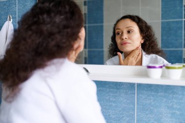 Çok ırklı 35 'li yaşlarda, genç esmer, beyaz bornozlu güzel bir kadın yaşlanma ve kırışıklıkların ilk belirtileri için aynada cildini inceliyor, nemlendirici sürüyor ve yüzüne masaj yapıyor..