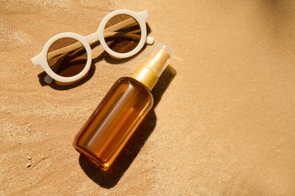Top view. Minimalist still life of unbranded dark glass bottle with SPF oil or sunscreen body lotion, sunglasses on golden sand beach background. Summer cosmetic concept. Copy ad space for text