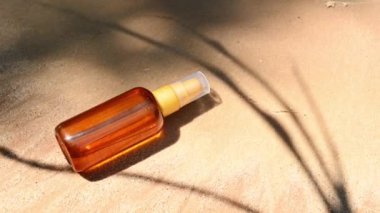 Minimalist still life of unbranded dark glass bottle with SPF oil or sunscreen body lotion on golden sand beach background with shadow from palm leaves. Summer cosmetic concept. Copy ad space for text