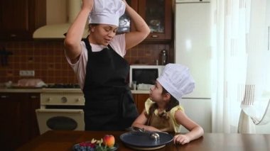 Pleasant multiethnic woman and her adorable little daughter, wearing a chef uniform talking together while cooking a sweet dessert in the home kitchen interior.