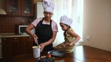 Charming multi-ethnic woman in a white chef hat and black apron, laying a frozen ball of delicious mango ice cream sorbet, for her daughter- adorable baby girl, sitting on a wooden kitchen table