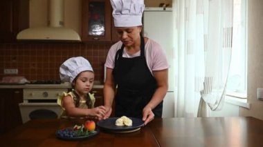 Inspired little chef, Caucasian child, cute girl in apron and chef hat, helps her mom to serve delicious homemade sweet dessert - frozen mango ice cream sorbet balls, cooking together at home kitchen