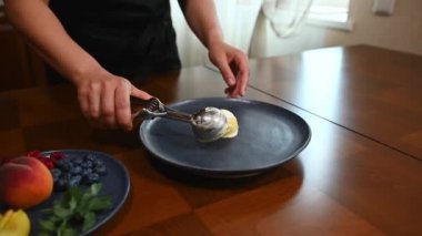 Close-up. Unrecognizable housewife, in a black chef apron places frozen balls of margo sorbet with a metal scoop on a plate and decorates with slices of fresh juicy and ripe peaches in home kitchen