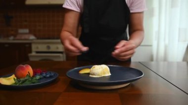 Close-up of unrecognizable housewife in a black chef apron, decorating balls of mango sorbet melting on a navy plate with slices of fresh ripe peach, blueberries and lemon basil leaves at home kitchen