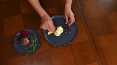 Top view. Chef hands, decorating frozen balls of mango sorbet melting on a navy ceramic plate with slices of fresh ripe peach, blueberries and lemon basil leaves at home kitchen. Healty raw vegan food