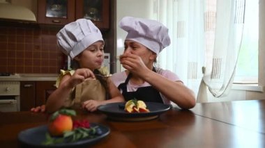 Loving mom with her daughter, in chef caps and aprons, eating delicious refreshing vegan mango sorbet at home kitchen. Charming woman feeding her beautiful baby girl, enjoying a tasty snack together