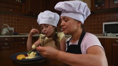 Cheerful mom and daughter wearing chef caps and aprons, enjoy healthy sweet snack together, sit at table and eat frozen mango ice cream balls at home kitchen. Woman and little girl taste vegan sorbet