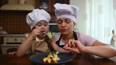 Adorable Caucasian child, little girl in chef cap and apron, eats delicious ice cream, topped with mango, passion fruits, peaches and wild berries, with her loving mother in the home kitchen interior