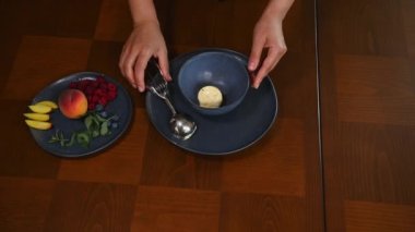 View from above of chef hands, using metal scoop for ice cream, placing mango sorbet balls on a blue navy bowl and decorating with berries and lemon basil leaves. Homemade raw vegan healthy dessert