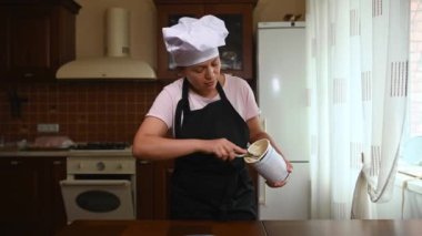 Multiethnic woman, housewife in white chef cap and black kitchen apron extracting mango sorbet balls with metal ice cream spoon while preparing sweet dessert in home wooden kitchen interior