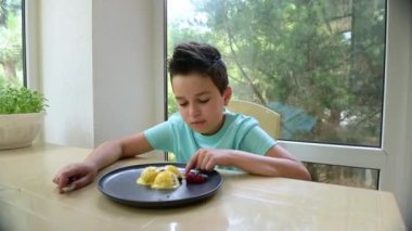 Adorable Caucasian ten-year-old boy enjoys the taste of a refreshing mango ice cream sorbet, shaped like balls and garnished with berries and fresh peach slices, while sitting on a summer veranda