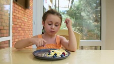Cute baby girl with two pigtails, wearing orange top, sitting on summer terrace, eats delicious sweet dessert- balls of mango passion fruit ice cream sorbet, topped with fresh sliced peach and berries