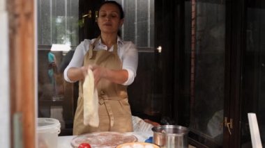 Inspired pleasant multiethnic middle-aged woman, a housewife in beige chef apron, tossing up the ready yeast dough while preparing bread or pizza, in old wooden rustic kitchen of her summer cottage