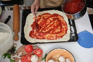 View from above. Housewife in beige chefs apron spreading freshly made tomato sauce on rolled out dough in pizza pan. Fresh organic ingredients on a kitchen table. Cooking. Culinary. Italian cuisine