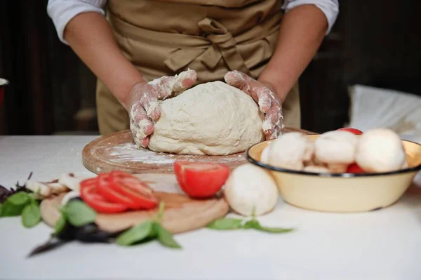 Details: Hands of a chef, wearing beige apron, with a raising yeast dough and fresh pizza ingredients: mushroom champignons, ripe organic tomatoes and basil leaves on the table on blurred foreground