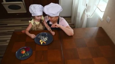 Top view. Beautiful loving mother, multiethnic young woman in chef cap and apron, feeding her adorable little daughter with a slice of ripe juicy peach while the girl finishes her delicious ice cream
