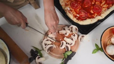 Details: Chefs hands slicing fresh mushroom champignons on a wooden chopping board, next to the pizza pan, organic vegetables and fragrant culinary leaves on a white table in rustic kitchen. Top view