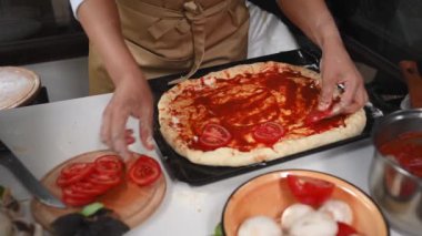 Close-up Hands of pizzaiolo, chef wearing kitchen apron, adding slices of fresh organic tomatoes on the rolled out dough, while preparing delicious Italian pizza in rustic kitchen. Restaurant business